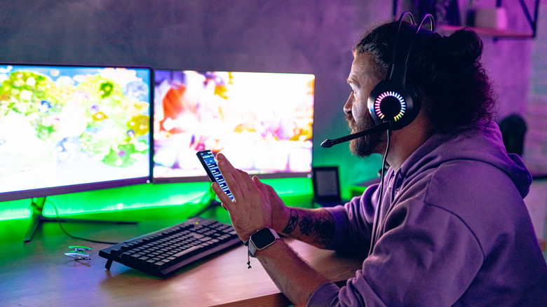 헤드폰을 착용하고 모니터 2개가 있는 책상에 앉아 스마트폰을 사용하는 사람.