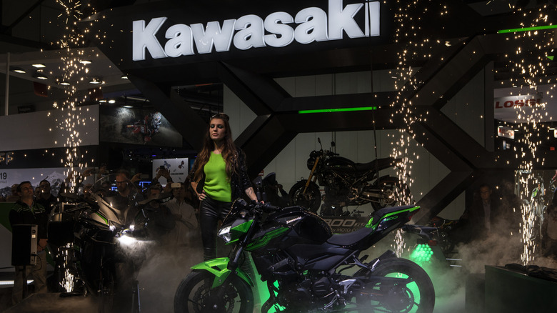 Kawasaki Versys 1000 오토바이가 조명이 켜진 Kawasaki 로고 아래 어둠 속에서 쇼에 모습을 드러내고 있습니다.