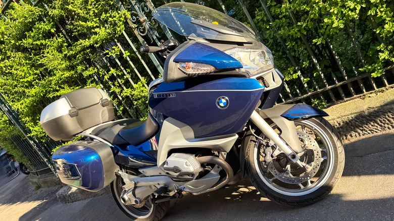 2012년에 제작된 파란색과 은색 BMW R1200RT 오토바이가 나뭇잎을 배경으로 도로변에 주차되어 있습니다.