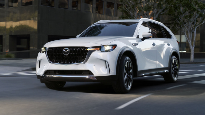 도시에서 빠른 속도로 코너링하는 Mazda CX-90 PHEV, 전면 3/4 뷰, 흰색 외관