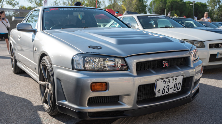 JDM 자동차 쇼에서 다른 JDM 자동차 옆에 주차된 회색 R34 Nissan Skyline.