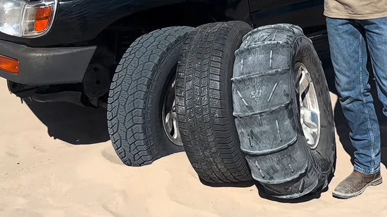 모래 위에 주차된 낡고 검은색 Toyota SUV. 이미 마모된 일반 전지형 타이어 옆에 두 개의 서로 다른 타이어 오타가 보입니다.