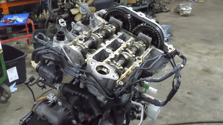 분해된 Ford 1.0리터 EcoBoost 엔진이 밸브 트레인, 캠축 및 타이밍 구성 요소가 작업장 조명 아래 완전히 노출된 채 분주한 작업장 내부 스탠드에 놓여 있습니다.