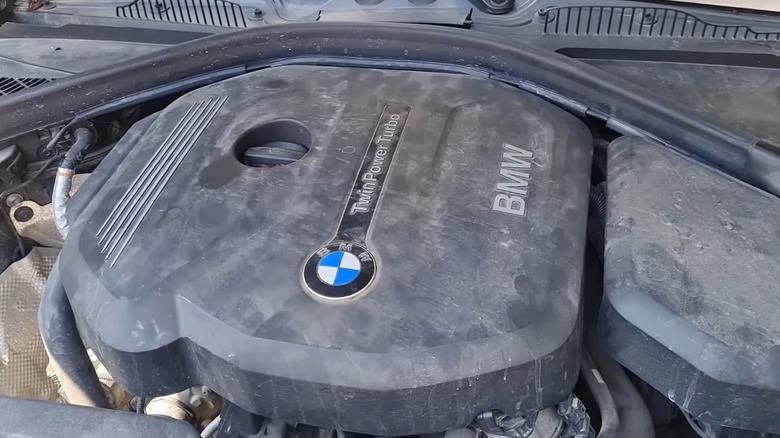 먼지가 쌓인 BMW B38 TwinPower Turbo 엔진 커버가 열린 후드 아래에 놓여 있습니다.
