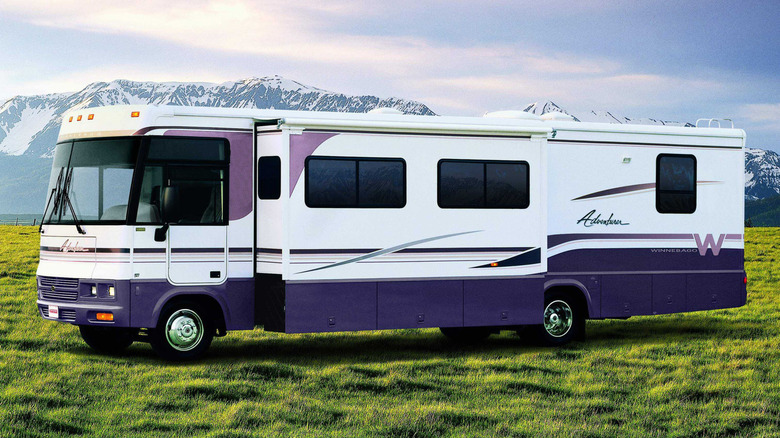 보라색 액센트가 있는 크고 현대적인 Winnebago와 눈 덮인 산을 배경으로 풀밭에 앉아 있는 미끄럼틀