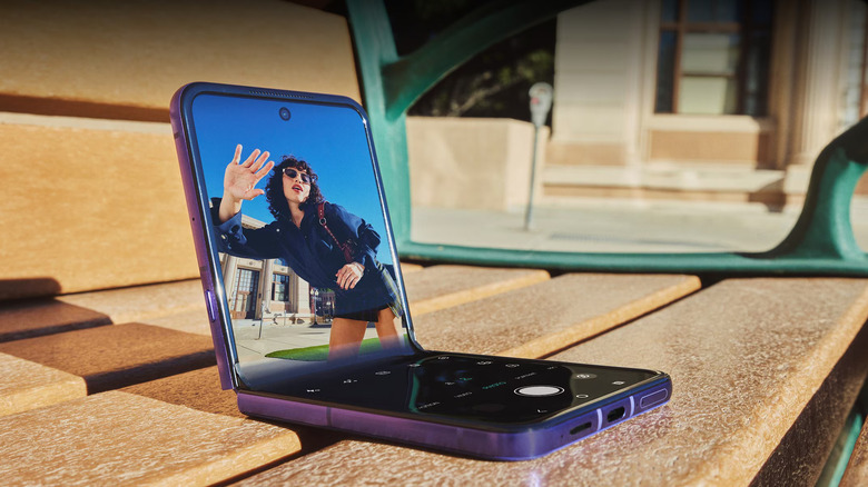 2026년형 모토로라 레이저 울트라(Motorola Razr Ultra)가 나무 벤치에 반쯤 접혀 있고 화면에 나타난 카메라를 향해 손을 흔드는 사람