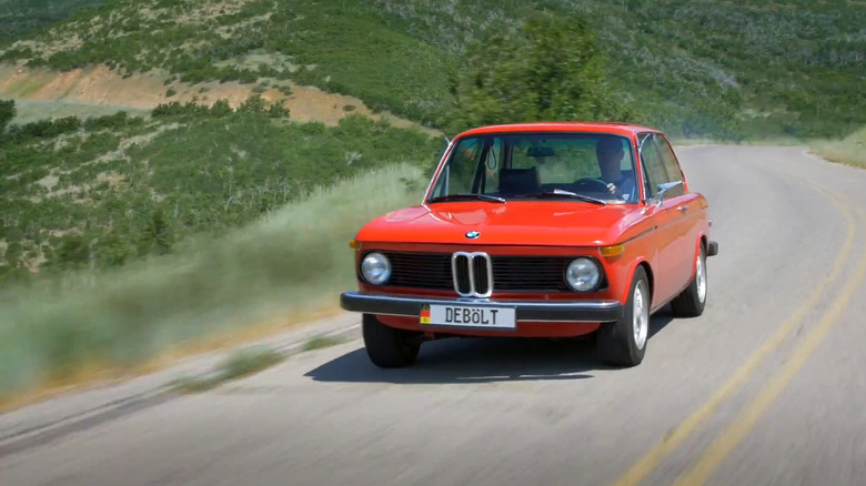 빨간색 1976년형 BMW 2002 터보 자동차가 뒤쪽에 계곡이 있는 곡선 산악 도로를 빠른 속도로 이동하고 있습니다.